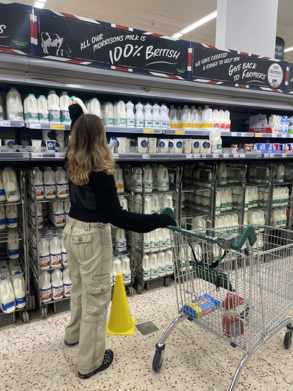 a young woman in a supermarket, she is wearing beige trousers and a black top. she is reaching up for some milk, and has a trolley with her. the trolley is mostly empty except for some biscuits and bread.