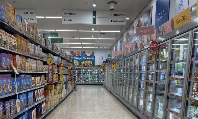 an empty supermarket aisle. on the left side there is a selection of cereals and on the right side there is a number of freezers containing ice cubes and ice cream.