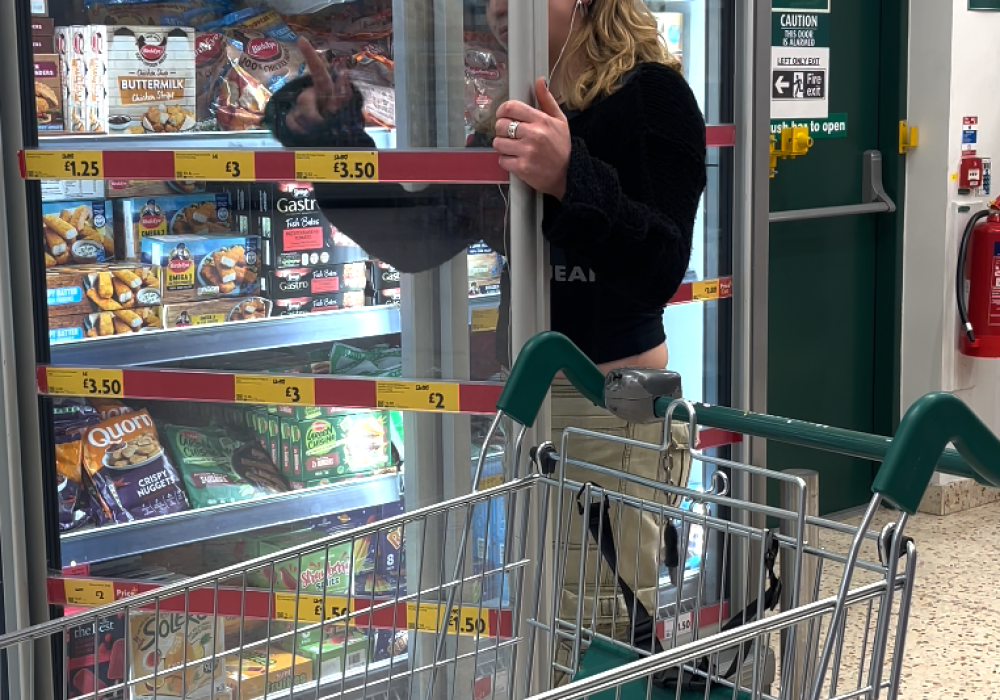 a young woman in a supermarket, she is wearing beige trousers and a black top. she trying to clean the inside of a freezer door with her jumper.