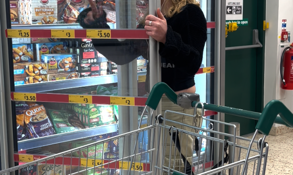 a young woman in a supermarket, she is wearing beige trousers and a black top. she trying to clean the inside of a freezer door with her jumper.