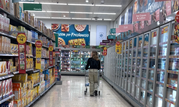 a supermarket aisle for cereals and frozen foods. there is only one young woman walking away from the camera. she is wearing beige trousers and a black top. she is pushing a trolley.