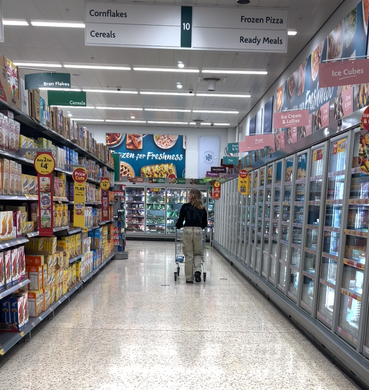 a supermarket aisle for cereals and frozen foods. there is only one young woman walking away from the camera. she is wearing beige trousers and a black top. she is pushing a trolley.