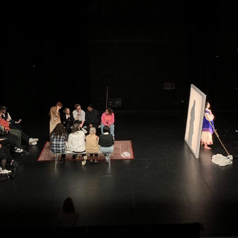 image of a performance work happening in a black box studio. there is a crowd sat on some cushions, looking at a woman stood behind a white screen.