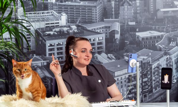 A receptionist wearing a spa uniform and headset; an orange cat is on the desk; a mobile phone with a candle emoji is also on the desk; a black and white backdrop of Leeds is behind the receptionist