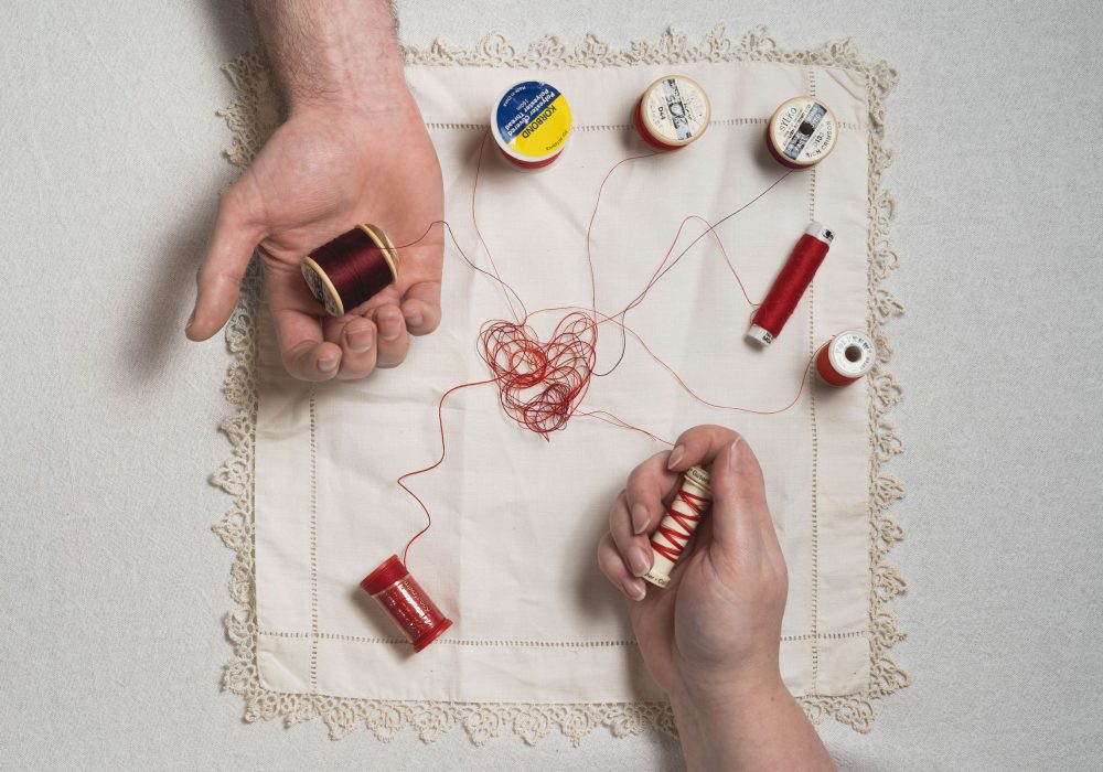 Two hands hold threads resting on an embroidered heart on a square of fabric