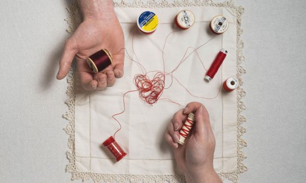 Two hands hold threads resting on an embroidered heart on a square of fabric