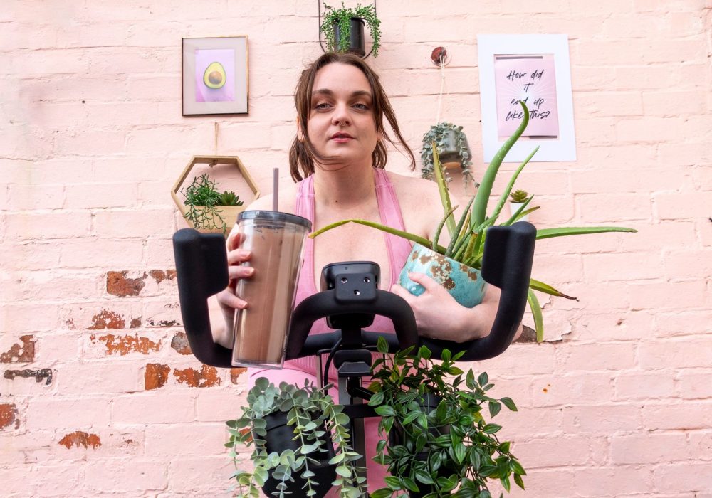 Lydia in milennial pink on a spin bike clutches a smoothie and a pot plant