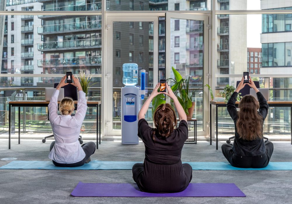 In an office setting, three people sit on yoga mats raising their phones above their heads