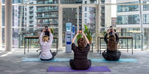 In an office setting, three people sit on yoga mats raising their phones above their heads