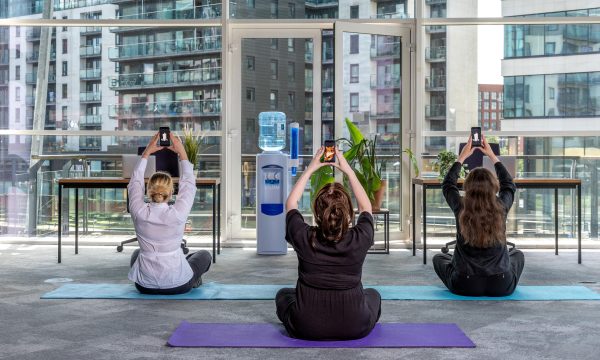 In an office setting, three people sit on yoga mats raising their phones above their heads