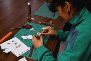 A person wearing a jade coloured jacket is holding a paper model of a house in both hands and is sitting a dark wooden table with a pair of scissors, cutting board & paper scraps on the surface